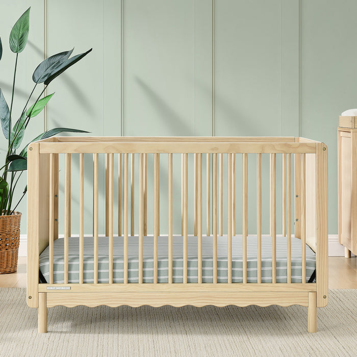 A William 4-in-1 Convertible Crib in beige color with scallop detailing, round spindles, and a light-colored dresser and chair in the background in a nursery room. 0-William 4-in-1 Convertible Crib - Greenguard Gold Certified