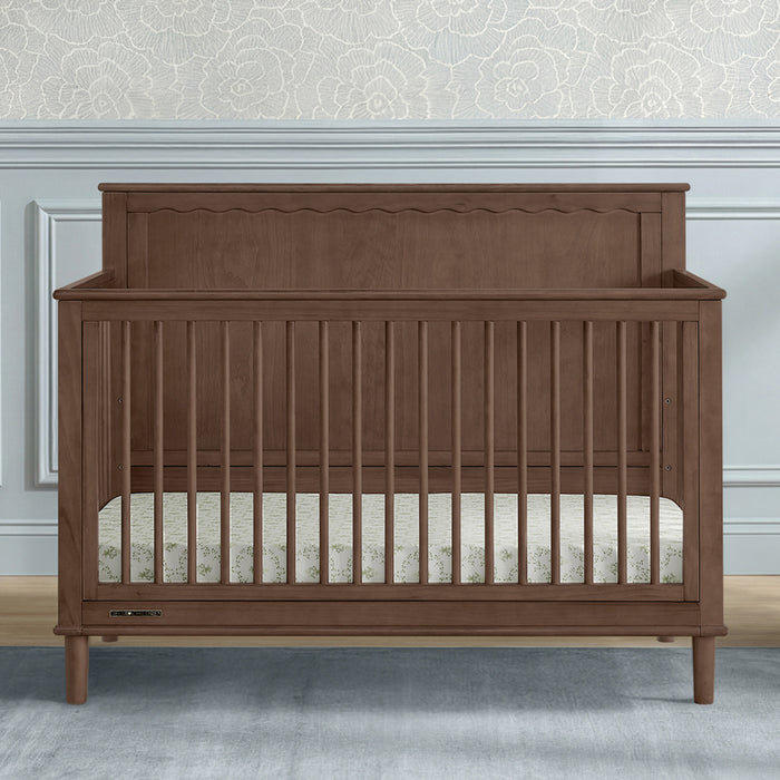 A wooden convertible crib with a warm brown finish, featuring vertical slats, a decorative headboard, and a mattress inside, set against a light gray wall with wainscoting and patterned wallpaper. 1-Penelope 6-in-1 Convertible Crib