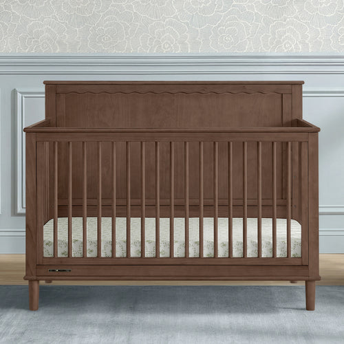 A wooden convertible crib with a warm brown finish, featuring vertical slats, a decorative headboard, and a mattress inside, set against a light gray wall with wainscoting and patterned wallpaper.