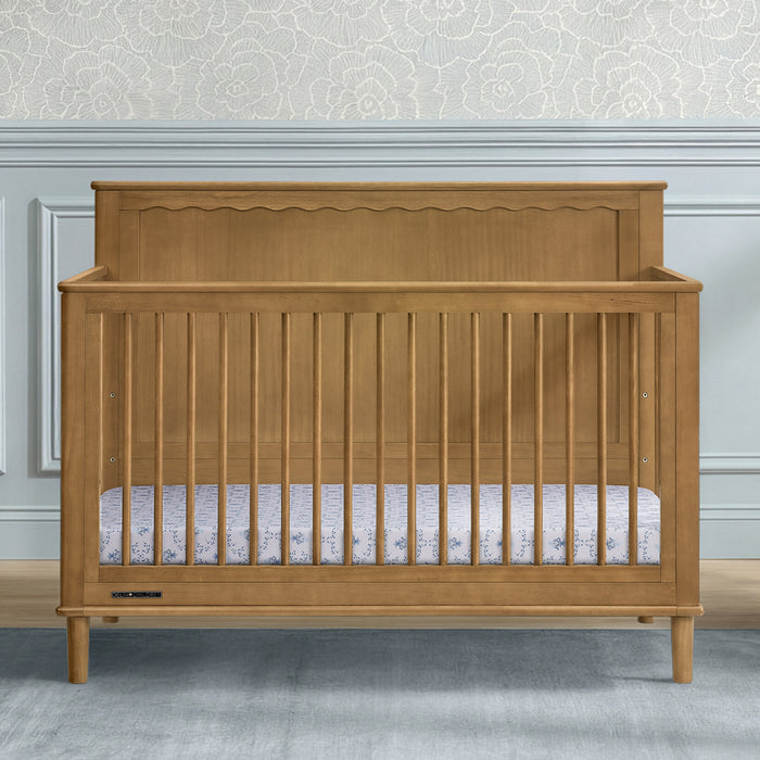 A wooden baby crib with a warm honey-oak finish, featuring a decorative scalloped headboard and vertical slats, shown in a styled nursery setting. 1-Penelope 6-in-1 Convertible Crib