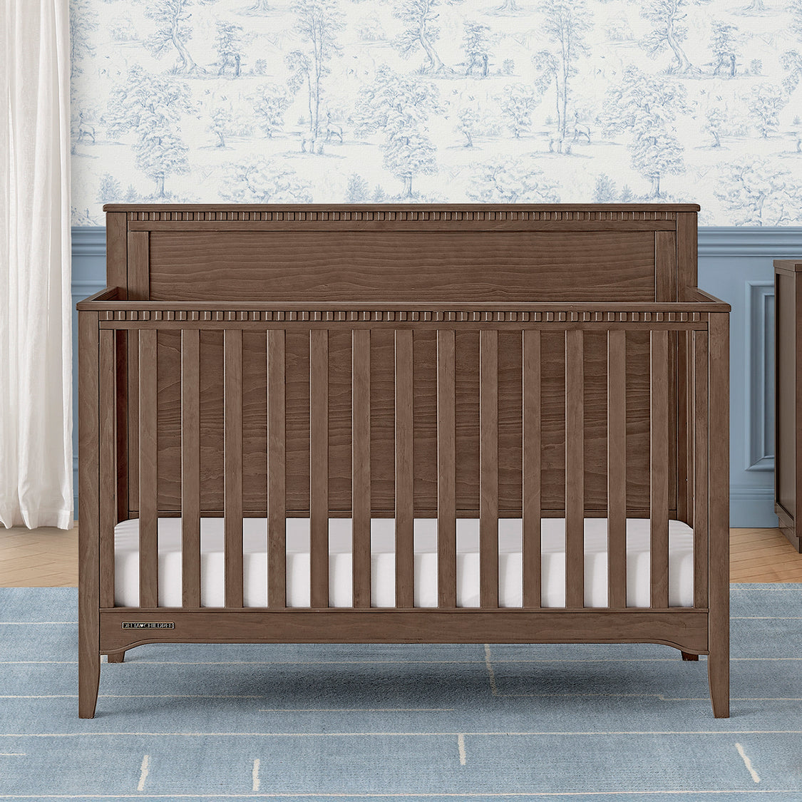 Wooden crib with white mattress in a room with floral wallpaper and gray walls.