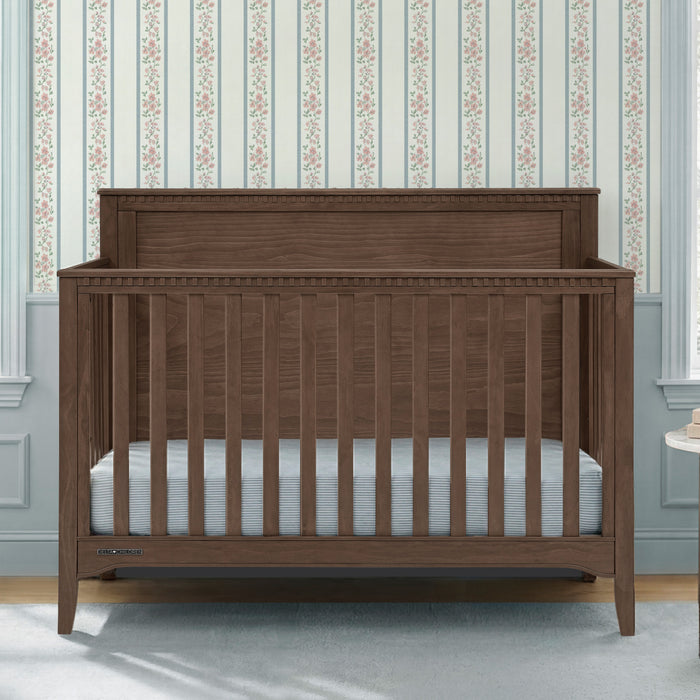 A dark brown wooden convertible crib with vertical slats and a high headboard, shown with a light blue striped mattress in a nursery setting with floral wallpaper. 3-Kendall 6-in-1 Convertible Crib