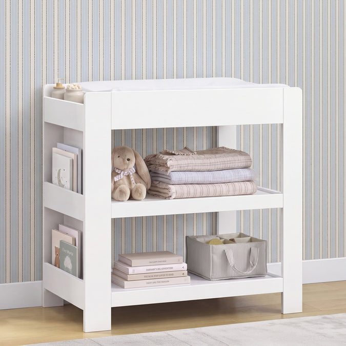 White wooden changing table with open shelves, styled with folded blankets, a teddy bear, books, and a storage basket against a striped wallpaper background. 6-Emmie Changing Table with Bookcase Storage