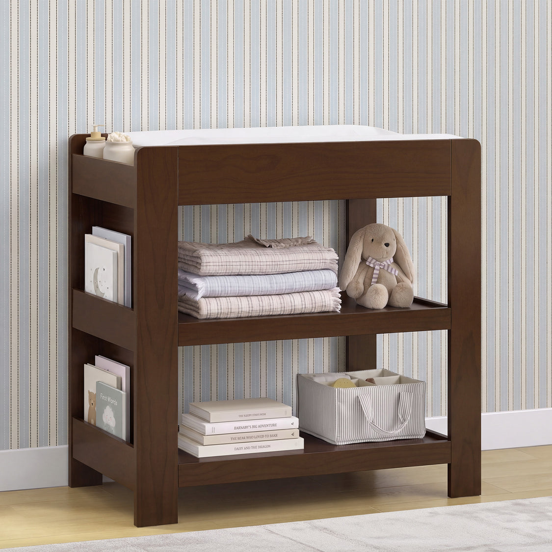 A dark brown wooden baby changing table with open shelves, styled with folded blankets, books, a stuffed bunny, and a storage basket against a striped wallpaper background.