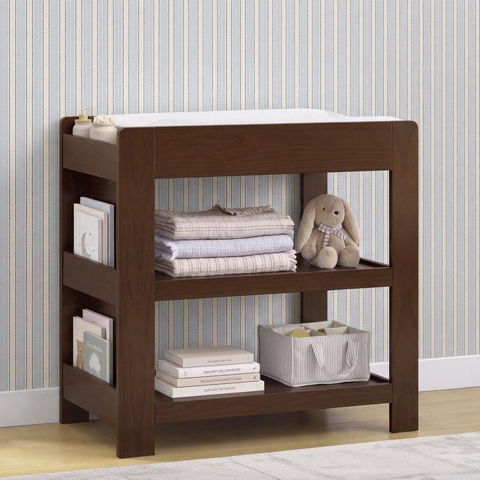 A dark brown wooden baby changing table with open shelves, styled with folded blankets, books, a stuffed bunny, and a storage basket against a striped wallpaper background. 12-Emmie Changing Table with Bookcase Storage