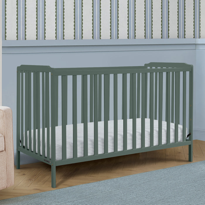 A sage green convertible crib with vertical slats, shown with a white mattress in a styled nursery setting with a light blue wall, wood floor, and rug. 3-Emmie 4-in-1 Convertible Crib