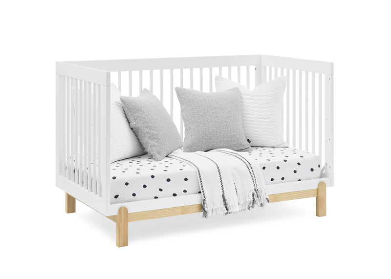 A white wooden crib with a natural wood frame base, styled with three gray and white pillows, a white mattress with black polka dots, and a light gray throw blanket, set against a plain white background.