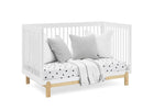 A white wooden crib with a natural wood frame base, styled with three gray and white pillows, a white mattress with black polka dots, and a light gray throw blanket, set against a plain white background. 12-Poppy 4-in-1 Convertible Crib