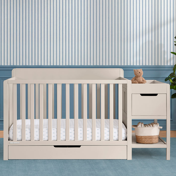 Beige crib with white bedding in a room with striped wallpaper 3-Maya 5-in-1 Convertible Crib and Changer with Storage
