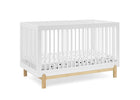 A white wooden crib with vertical slats and light natural wood legs, shown on a clean white background. A white mattress with small black polka dots is visible inside. 23-Poppy 4-in-1 Convertible Crib