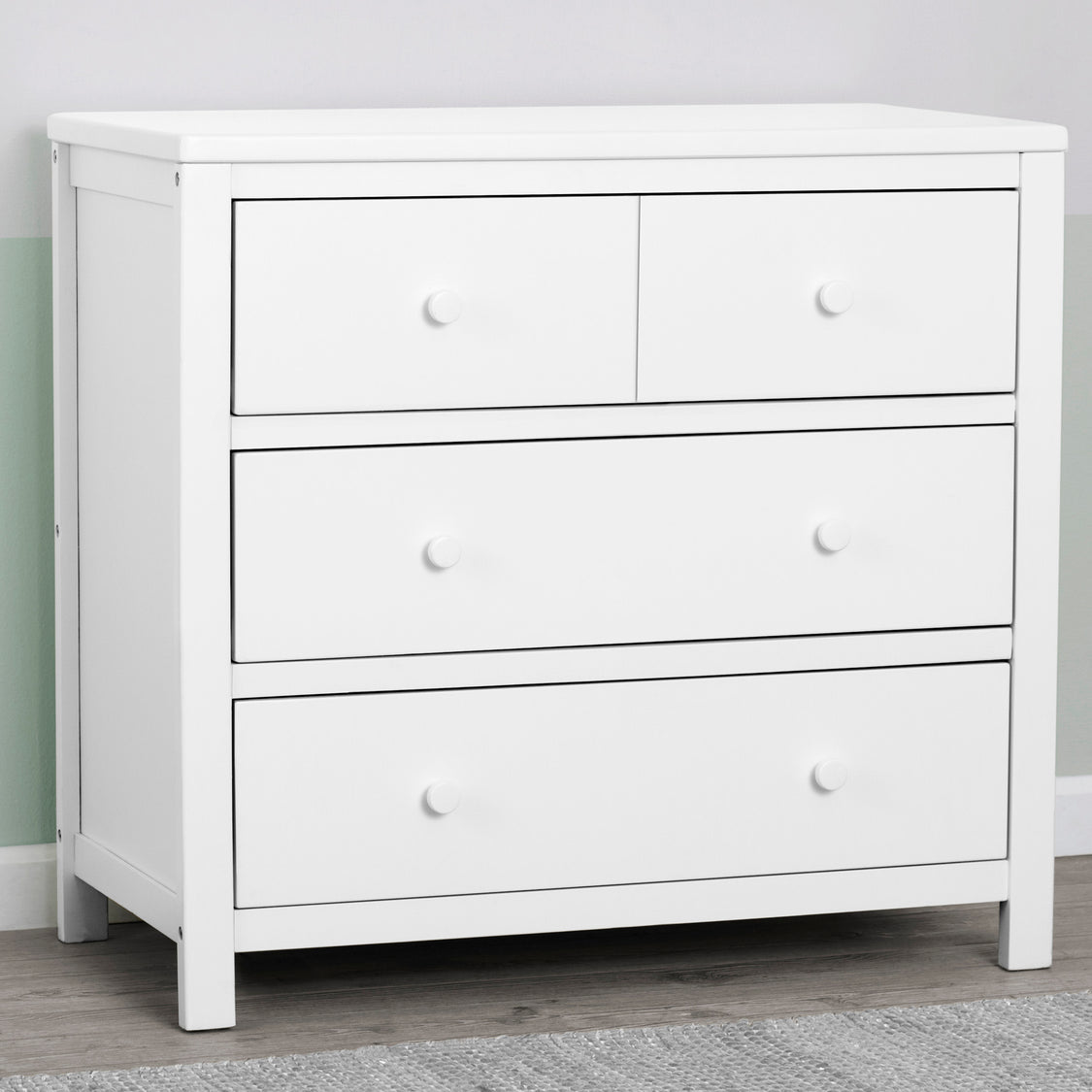 A white 3 drawer dresser with a closed drawer system, positioned against a light-colored wall on a grey floor.
