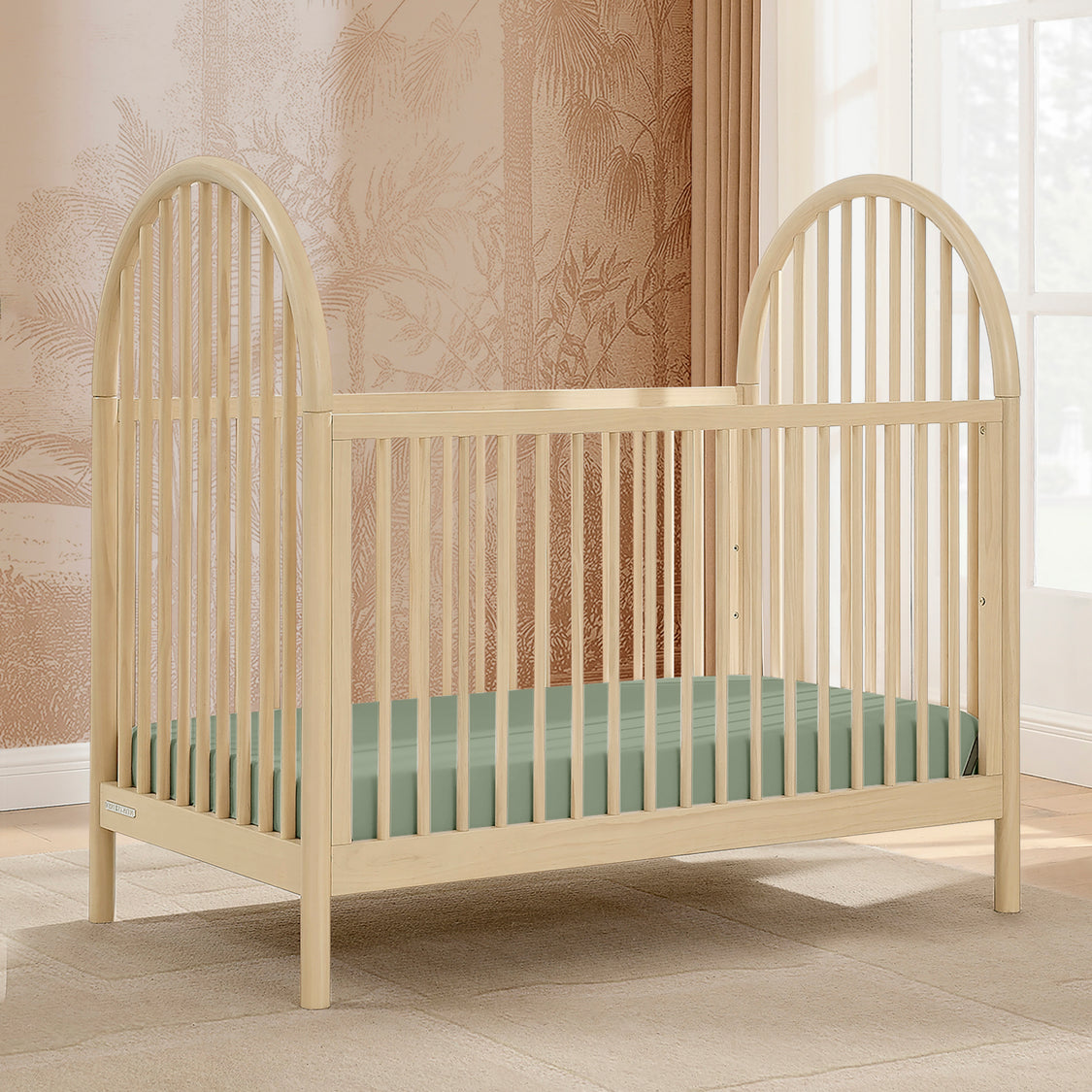 Beige crib with a green mattress in a room with patterned wallpaper and a window.