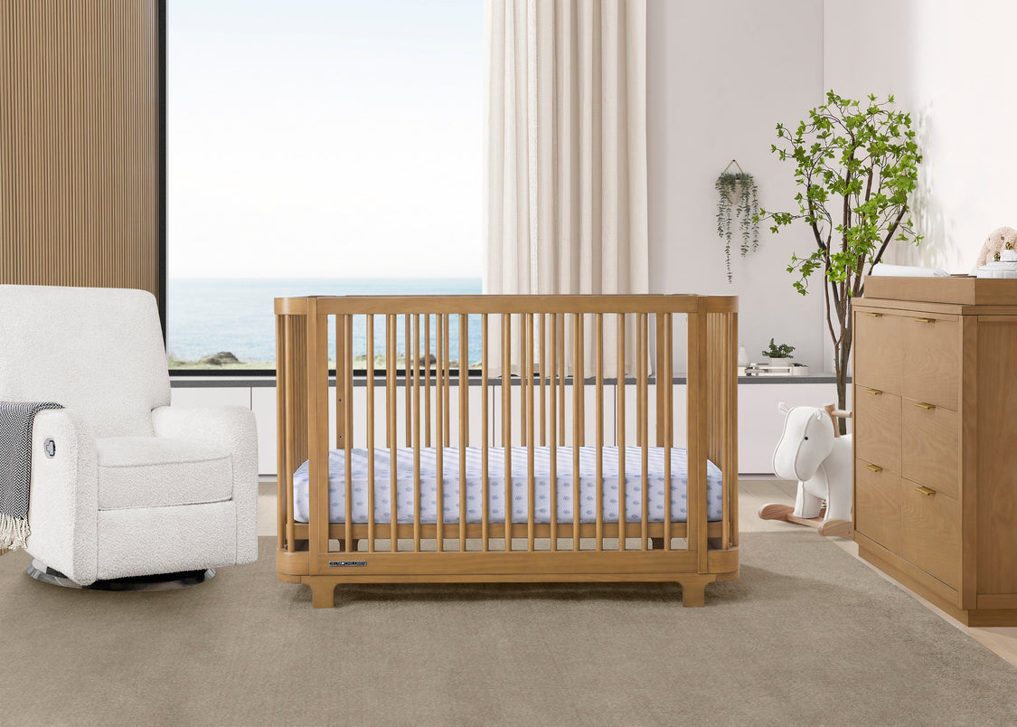 A brown Nest 4-in-1 Convertible Crib by Delta Children in a modern nursery room, with a white mattress on a white floor, alongside a white chair and a wooden dresser.
