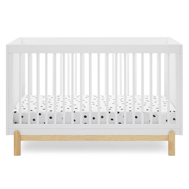 A modern white baby crib with vertical slats and a natural light wood base, shown with a white mattress fitted sheet featuring small black polka dots, on a clean white background.