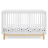A modern white baby crib with vertical slats and a natural light wood base, shown with a white mattress fitted sheet featuring small black polka dots, on a clean white background. 15-Poppy 4-in-1 Convertible Crib