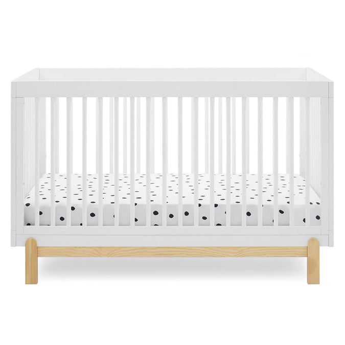 A modern white baby crib with vertical slats and a natural light wood base, shown with a white mattress fitted sheet featuring small black polka dots, on a clean white background. 15-Poppy 4-in-1 Convertible Crib