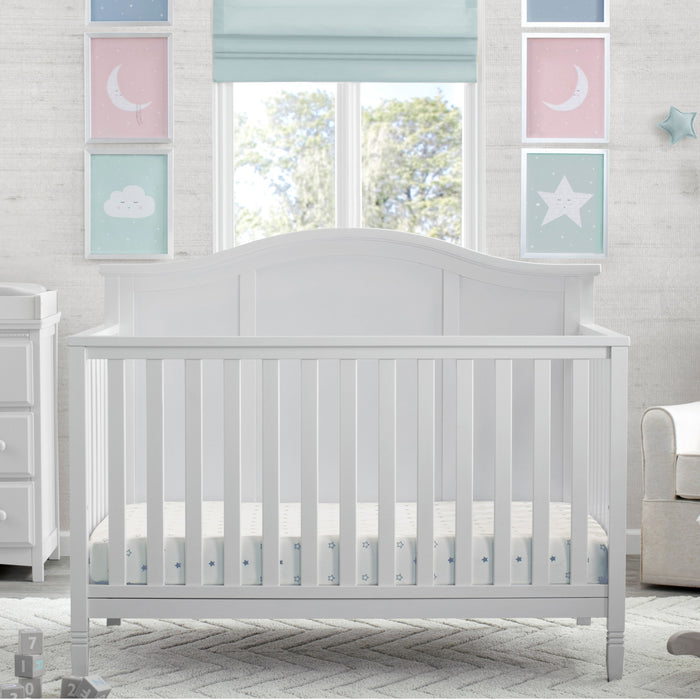 A white 5-in-1 convertible crib with a headboard, situated in a nursery room with decorative wall art, curtains, and furniture. 2-Madrid 5-in-1 Convertible Crib
